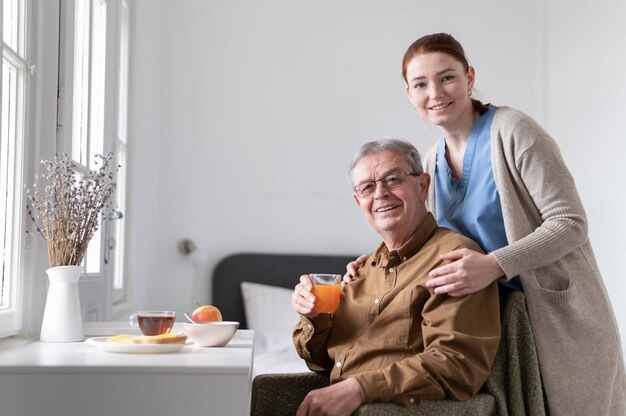 Assisted Living for Seniors: Enjoy a Safe, Fulfilling Life 2 Caregiver standing beside a smiling senior man holding juice, offering support in a bright home with breakfast on the table.