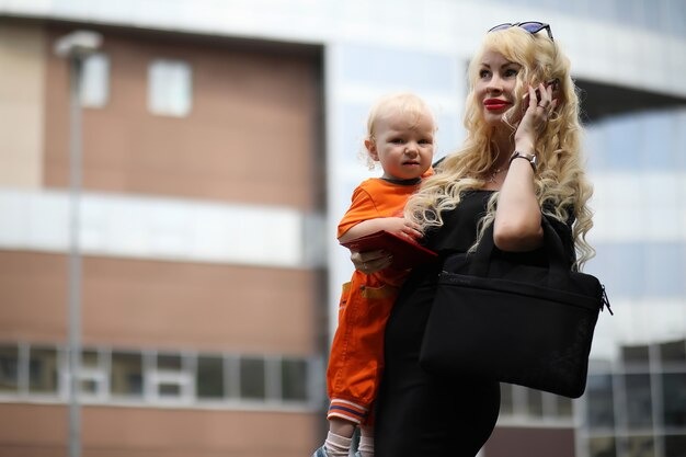 A blonde woman in a black outfit holding a baby dressed in orange while talking on her phone outside a modern building.