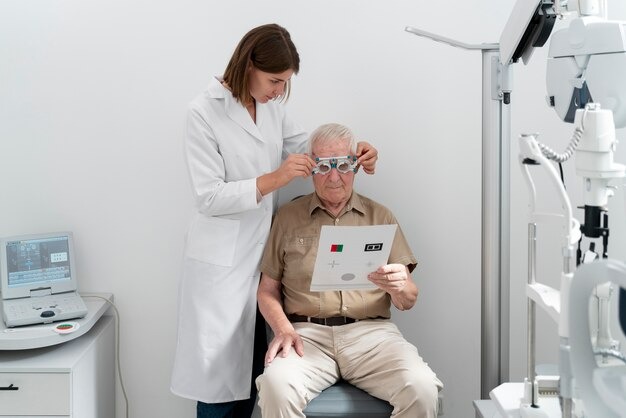 A female eye doctor adjusts trial lenses on an elderly man while he reads an eye chart in a clinic exam room.