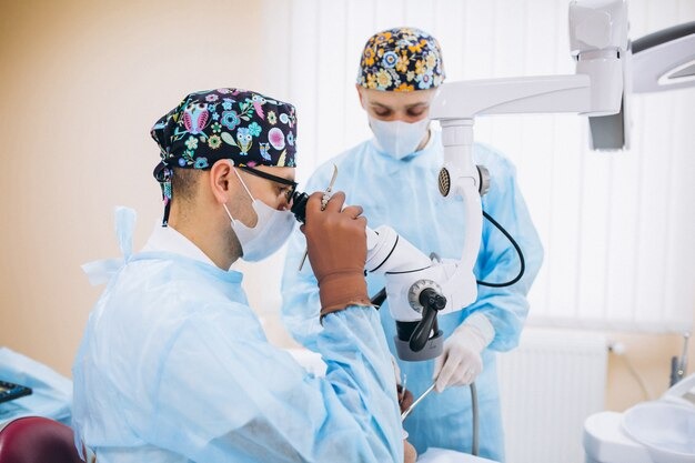 Two eye surgeons wearing masks and surgical caps use a microscope while performing an eye procedure in an operating room.

