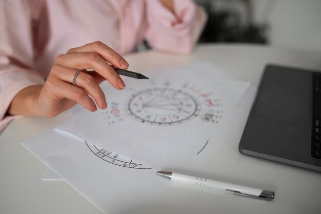 Close-up of a person holding a pen over printed astrological charts next to a laptop.