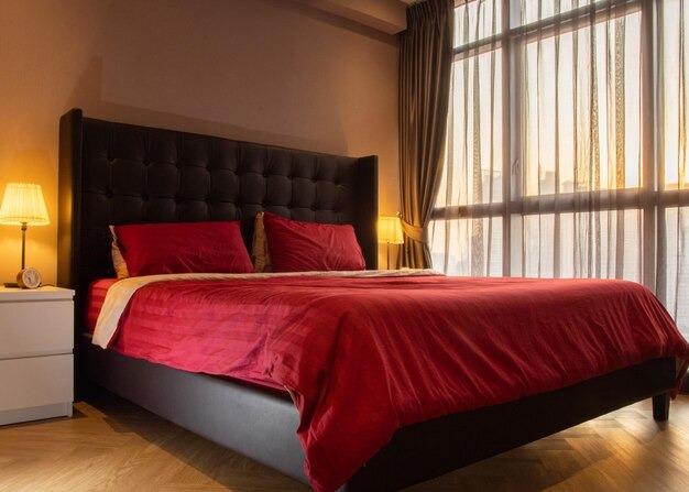 Alt text: Modern bedroom with red bedding, dark upholstered bed, bedside lamps, and large window with sheer curtains.