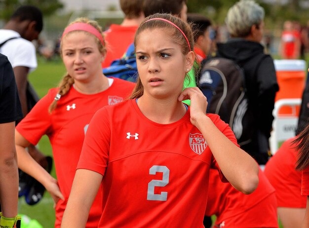 Female soccer players in red uniforms on field; player wearing number 2 looks concerned during game break.