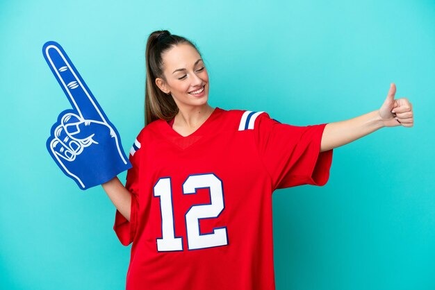 Smiling woman in red sports jersey holding foam finger and giving thumbs up against blue background.